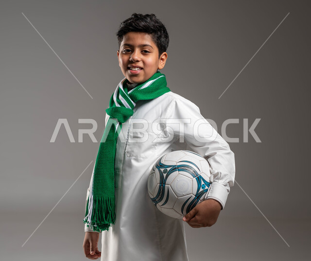 جاهز للتحدي والفوز يرتدي الثوب التقليدي ووشاح أخضر يحمل بيده كرة قدم بتعابير الفرح والسرور والحماس للمنافسة، بورتريه مقرّب لولد عربي خليجي سعودي ، تشجيع الفريق السعودي، النظر إلى الكاميرا بروح العزيمة على الانتصار، خلفية رمادية