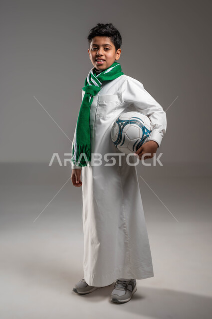 جاهز للتحدي والفوز، بورتريه لولد عربي خليجي سعودي يرتدي الثوب التقليدي ووشاح أخضر يحمل بيده كرة قدم بتعابير الفرح والسرور والحماس للمنافسة، تشجيع الفريق السعودي، النظر إلى الكاميرا بروح العزيمة على الانتصار، صورة الجسم بالطول الكامل، خلفية رمادية