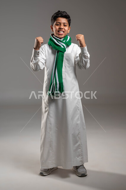 Looking at the camera with gestures of pleasure and joy at achieving victory and triumph, portrait of a Saudi Gulf Arab boy wearing a traditional dress and a green scarf, raising his hands up with an expression of enthusiasm, cheering for the Saudi team, full-length body portrait, gray background