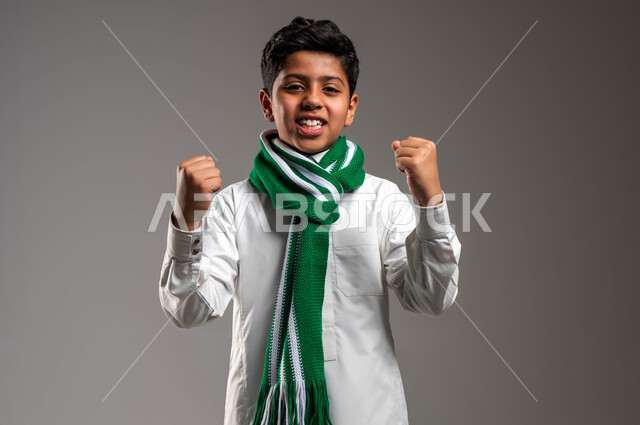 تشجيع الفريق السعودي، بورتريه لولد عربي خليجي سعودي يرتدي الثوب التقليدي والوشاح الأخضر وينظر الى الكاميرا بايماءات السرور، رفع اليدين للأعلى بكفين مقبوضتين بحركات الحماس، تعابير الفرح بتحقيق الفوز والانتصار، خلفية رماديه