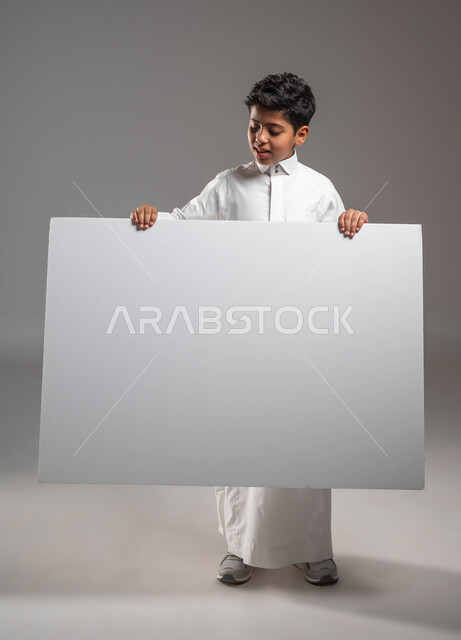 النظر إلى لوحة مربعة بإيماءات السرور، استخدام لوحات تعبيرية توضيحية، موك أب مربع أبيض, لوح إعلاني فارغ، بورتريه لولد عربي خليجي سعودي مبتسم  يرتدي الثوب التقليدي يقف بإستقامة ويمسك بيده لوحة مربعة بيضاء، خلفية رمادية