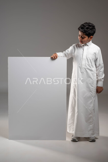 استخدام لوحات تعبيرية توضيحية، موك أب مربع أبيض, لوح إعلاني فارغ، النظر إلى لوحة مربعة بإيماءات السرور، بورتريه لولد عربي خليجي سعودي مبتسم  يرتدي الثوب التقليدي يقف بإستقامة ويمسك بيده لوحة مربعة بيضاء، صورة الجسم بالطول الكامل، خلفية رمادية