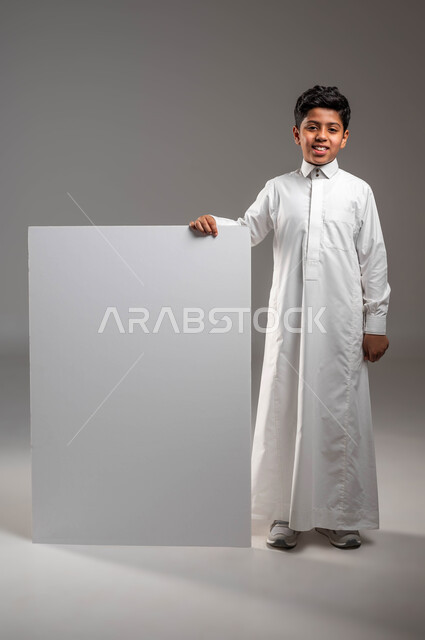 White square mockup, empty billboard, looking at the camera with happy gestures, using expressive illustrative paintings, portrait of a smiling Saudi Arabian Gulf boy wearing traditional dress, standing straight and holding a white square billboard in his hand, full-length body portrait, gray background