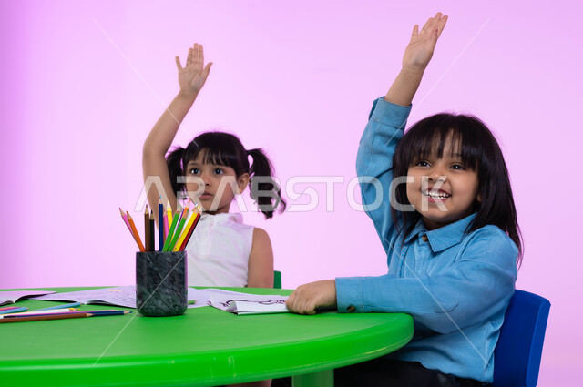 المشاركة برفع اليد أثناء الحصة، قرطاسية وأدوات مدرسية للأطفال، الاندماج والتركيز في الحصص والجلسات الفنية في الروضات السعودية، بورتريه مقرب لأطفال عرب خليجيين سعوديين يرتدون الزي الكاجوال يرفعون يديهم للأعلى، خلفية وردية