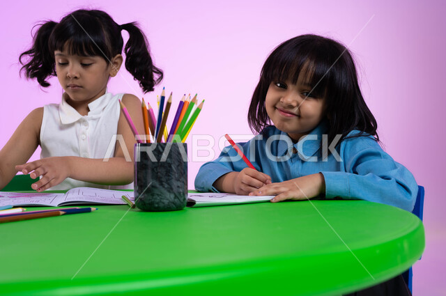 الجلسات والحصص التعليمية في الروضات السعودية، تنمية القدرات العقلية والذهنية بالمهارات و الأنشطة المختلفة، الاندماج والتركيز في الرسم والتلوين، بورتريه مقرب لأطفال عرب خليجيين سعوديين يرتدون الزي الكاجوال يقومون بنشاط طلاء الرسومات، خلفية وردية