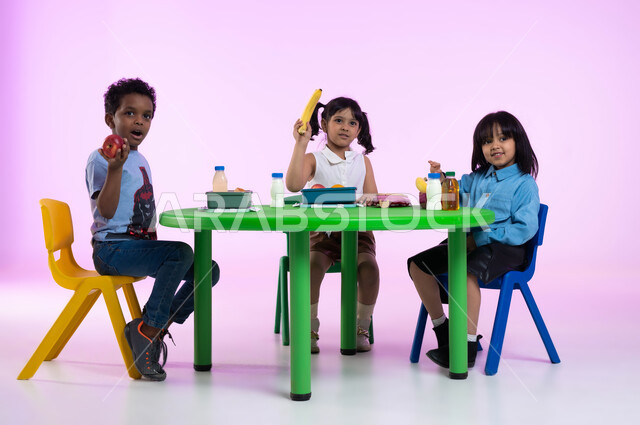 أنشطة لتعليم الأولاد في الروضة، مدارس وحضانات ومراكز ضيافة للأطفال، تعليم الأولاد في الروضة أسس الغذاء الصحي السليم لتقوية أجسامهم ونموها، بورتريه لأطفال عرب خليجيين سعوديين يرتدون الزي الكاجوال يتناولون الطعام الصحي وقت الفسحة المدرسية في إحدى رياض الأطفال في السعودية، خلفية وردية