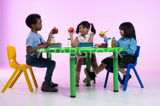 مدارس وحضانات ومراكز ضيافة للأطفال، تعليم الأولاد في الروضة أسس الغذاء الصحي السليم لتقوية أجسامهم ونموها، بورتريه لأطفال عرب خليجيين سعوديين يرتدون الزي الكاجوال يتناولون الطعام الصحي وقت الفسحة المدرسية في إحدى رياض الأطفال في السعودية، خلفية وردية