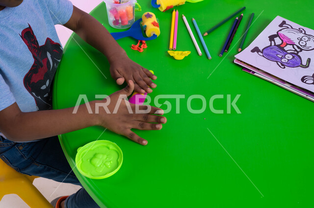 قرطاسية وأدوات مدرسية، متعة اللعب بمعجون التشكيل الملون، نشاطات فنية تساعد على تنمية الجانب الإبداعي والابتكاري لدى الأطفال، بورتريه علوي لطفل عربي خليجي سعودي يرتدي الزي الكاجوال يقوم باللعب بالصلصال في الروضة، خلفية وردية