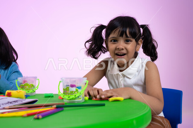 متعة اللعب بعجينة التشكيل الملونة، نشاطات فنية تساعد على تنمية الجانب الإبداعي والابتكاري لدى الأطفال، بورتريه لطفلة عربية خليجية سعودية ترتدي الزي الكاجوال تنظر إلى الكاميرا وتعلب بالصلصال، خلفية وردية.