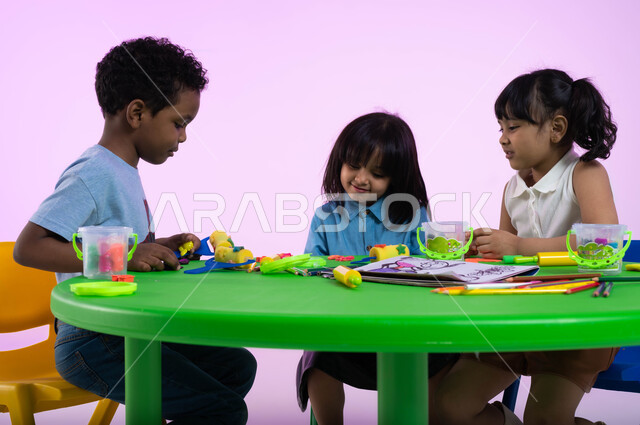 الاندماج والتركيز في تشكيل مجسمات وأشكال باستخدام العجينة الملونة, تعزيز الإبداع والمهارات الحركية والحسية الدقيقة عند الأولاد، بورتريه لأطفال عرب خليجيين سعوديين يبدعون بنشاط اللعب بالمعجون الملون في الروضة، خلفية وردية