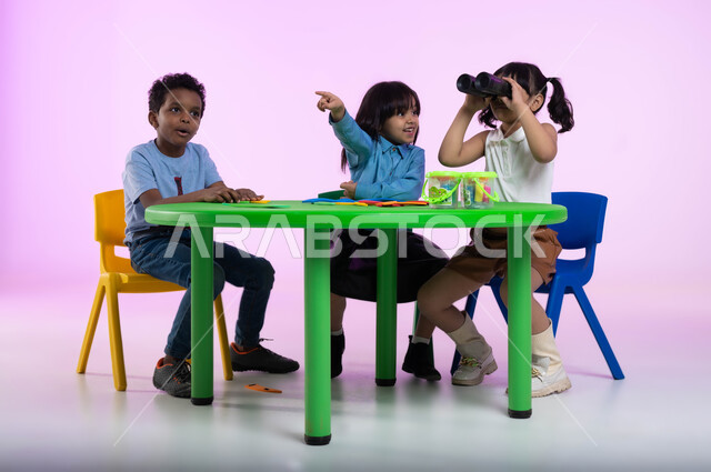 نظرة مستقبلية لدى الأولاد, إيماءات السعادة والدهشة، بورتريه لأطفال عرب خليجيين سعوديين يرتدون الزي الكاجوال يجلسون في الروضة، طفلة سعودية تحمل بيدها المنظار ثنائي العينين، الإشارة إلى شيء ما، حب الاستكشاف والمغامرة، خلفية وردية