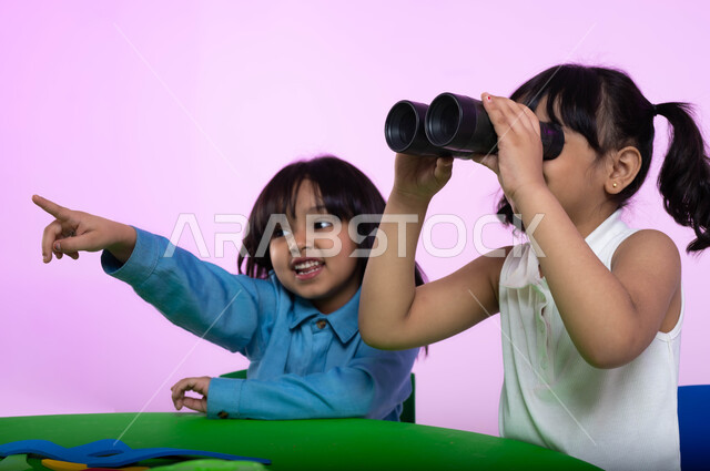 الإشارة إلى شيء ما، حب الاستكشاف والمغامرة، نظرة مستقبلية لدى الأولاد, بورتريه لطفلتين عربيتين خليجيتين سعوديتين يرتديان الزي الكاجوال، طفلة سعودية تحمل بيدها المنظار ثنائي العينين، إيماءات وجه تدل على السعادة والفرح، خلفية وردية.