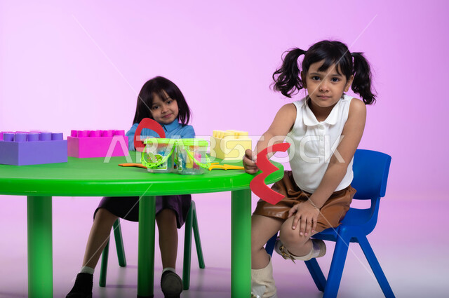 تعلم الأرقام والأعداد العربية وأشكالها، نشاطات ترفيهية وتعليمية للأطفال في الروضة، بوتريه لطفلتين عربيتين خليجيتين سعوديتين ترتديان الزي الكاجوال تنظران إلى الكاميرا بإيماءات تدل على السعادة والثقة بالنفس، خلفية وردية.