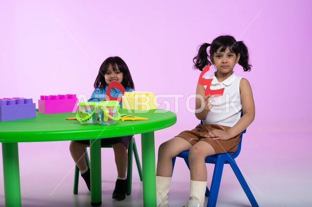 نشاطات تساعد على التطور الفكري، الجلسات والحصص الإبداعية في الروضات السعودية، إيماءات تدل على السعادة والمتعة، بورتريه لأطفال عرب سعوديين خليجيين يرتدون زي الكاجوال  يستمتعون بتركيب المكعبات، خلفية وردية.