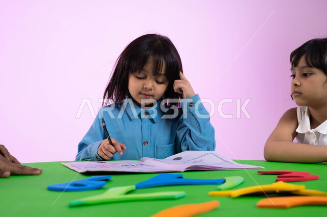 أنشطة تعليمية تساعد على تنمية القدرات العقلية والذهنية عند الأولاد، طفلة عربية خليجية سعودية تفكر وتحاول حل التمارين الحسابية في الروضة، بورتريه لأطفال عرب سعوديين خليجيين يرتدون الزي الكاجوال يتعاونون في حل الواجبات، خلفية وردية.