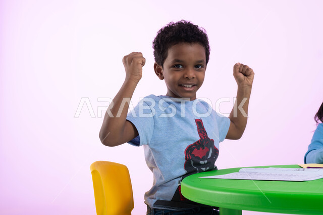 التشجيع والمنافسة والتحدي بين الأطفال في الروضة، بورتريه مقرب لطفل عربي خليجي سعودي يرتدي الزي الكاجوال يرفع يديه مضموم الكفين، إيماءات الحماس والشغف والسعادة بالفوز والإنتصار، النجاح والقوة وتحقيق الأهداف، خلفية وردية.