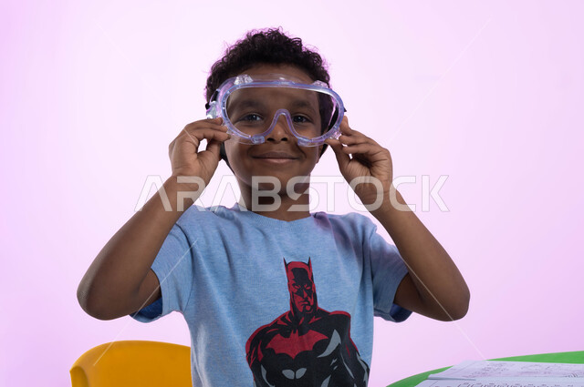 الحماس والشوق للغوص في الماء، أنشطة ترفيهية وتعليمية للأطفال، بورتريه مقرب لطفل عربي خليجي سعودي يرتدي الزي الكاجوال ونظارات السباحة للاستعداد والتحضير للسباحه، اللعب واللهو بأدوات المسبح، خلفية وردية.