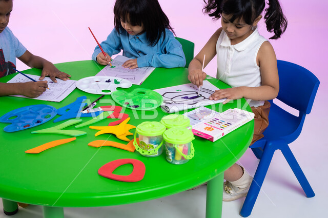 أنشطة ترفيهية وفنية في الروضة، تنمية المهارات والتعليم والإبداع، تقنيات الرسم لمرحلة ما قبل الدرسة، الحصص الابداعية في الروضات، بورتريه لأطفال عرب خليجيين سعوديين يرتدون الزي الكاجوال يبدعون بنشاط تلوين الرسومات بإيماءات وجه تدل على السعادة، خلفية وردية