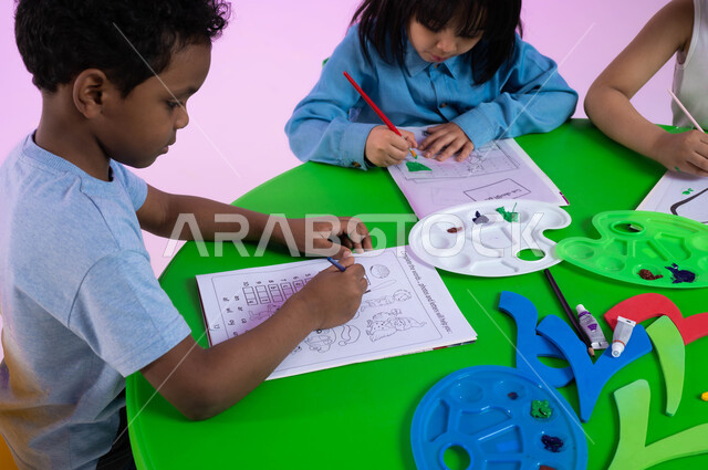 تقنيات الرسم لمرحلة ما قبل المدرسة، أنشطة ترفيهية وفنية في الروضة، تنمية المهارات والتعليم والإبداع، الحصص الابداعية في الروضات، بورتريه لأطفال عرب خليجيين سعوديين يرتدون الزي الكاجوال يبدعون بنشاط تلوين الرسومات بإيماءات وجه تدل على السعادة، خلفية وردية