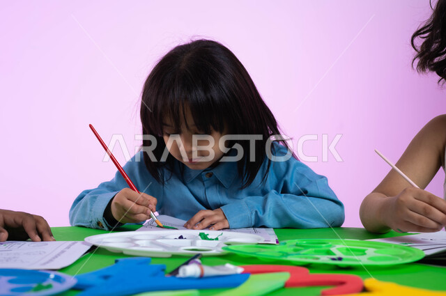 أنشطة ترفيهية وفنية في الروضة، تنمية المهارات والتعليم والإبداع، تقنيات الرسم لمرحلة ما قبل الدرسة، الحصص الابداعية في الروضات، بورتريه مقرب لطفلة عربية خليجية سعودية ترتدي الزي الكاجوال تبدع بنشاط تلوين الرسومات بإيماءات وجه تدل على السعادة، خلفية وردية