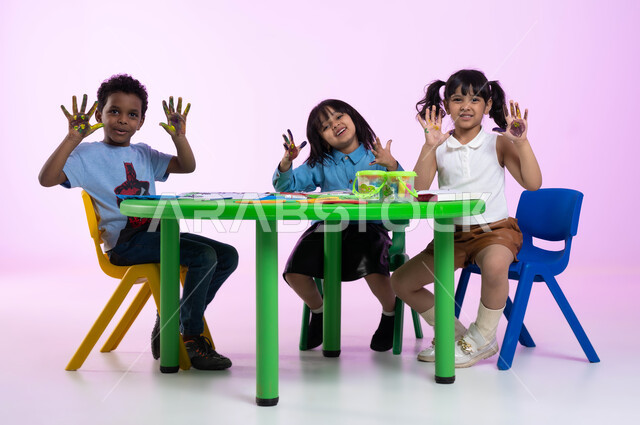 Watercolor palm printing, creative classes in kindergartens, activities that help intellectual development, drawing techniques for preschoolers, gestures indicating pleasure, close-up portrait of Saudi Arabian Gulf children wearing casual clothes showing their colorful hands, pink background