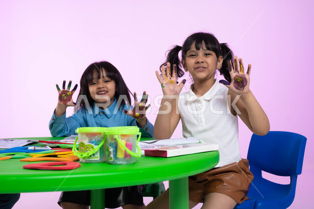 تقنيات الرسم لمرحلة ما قبل المدرسة، طباعة الألوان المائية بكف اليد، الحصص الإبداعية في الروضات، أنشطة تساعد على التطور الفكري، إيماءات تدل على المتعة، بورتريه مقرب لطفلتين عربيتين سعوديتين خليجيتين ترتديان الزي الكاجوال تظهران أيديهما الملونة، خلفية وردية