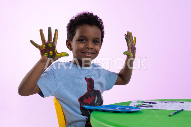 Creative classes in kindergartens, activities that help intellectual development, drawing techniques for pre-schoolers, gestures indicating pleasure, close-up portrait of a Saudi Arabian Gulf child wearing a casual outfit showing his colorful hands, watercolor palm print, pink background