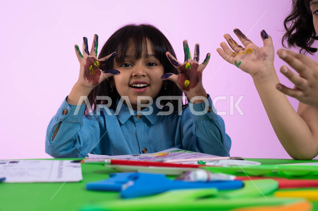 Activities that help intellectual development, creative classes in kindergartens, drawing techniques for pre-schoolers, gestures indicating pleasure, close-up portrait of a Saudi Arabian Gulf girl wearing a casual outfit showing her colorful hands, watercolor palm print, pink background