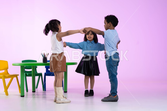 العاب تعليمية ترفيهية، قضاء أوقات ممتعة ومفيدة في الروضة، مفهوم دمج المتعة بالفائدة، تعليم وإبداع وتنمية مهارات في رياض الأطفال، بورتريه لأطفال عرب خليجيين سعوديين يرتدون الزي الكاجوال يلعبون ويمرحون، إيماءات تدل على الفرح والسعادة، صورة لكامل الجسم، خلفية وردية