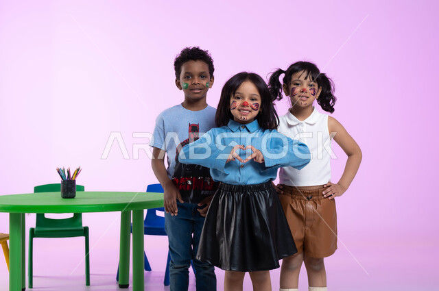 النظر إلى الكاميرا بتعابير تدل على السعادة، إيماءات يدين على شكل قلب، الشعور بالحب والإعجاب، الرسم على وجوه الأطفال بالالوان الصحية، قضاء أوقات ممتعة ومفيدة في الروضة، بورتريه لأطفال عرب خليجيين سعوديين يرتدون الزي الكاجوال، خلفية وردية