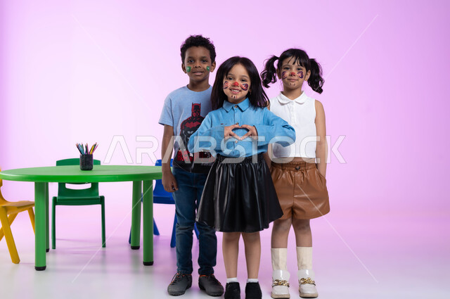 الشعور بالحب والإعجاب، الرسم على وجوه الأطفال بالالوان الصحية، قضاء أوقات ممتعة ومفيدة في الروضة، بورتريه لأطفال عرب خليجيين سعوديين يرتدون الزي الكاجوال ينظرون إلى الكاميرا بتعابير تدل على السعادة، إيماءات يدين على شكل قلب، صورة لكامل الجسم، خلفية وردية