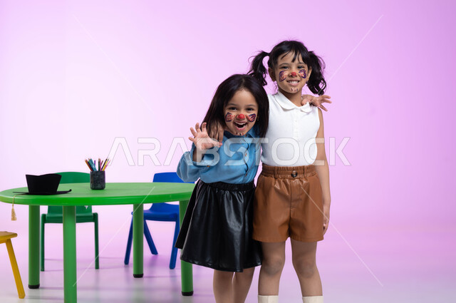 التظاهر والتمثيل بالحيوانات المفترسة، قضاء أوقات ممتعة بالقيام بأنشطة ترفيهية في الروضة، بورتريه لطفلتين عربيتين خليجيتين سعوديتين ترتديان الزي الكاجوال تلعبان وتمرحان، الرسم بألوان صحية على وجوه الأطفال، ايماءات تدل على الفرح والسعادة، خلفية وردية
