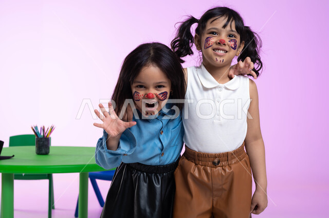 قضاء أوقات ممتعة بالقيام بأنشطة ترفيهية في الروضة، بورتريه لطفلتين عربيتين خليجيتين سعوديتين ترتديان الزي الكاجوال تلعبان وتمرحان، التظاهر والتمثيل بالحيوانات المفترسة،  الرسم بألوان صحية على وجوه الأطفال، ايماءات تدل على الفرح والسعادة، خلفية وردية