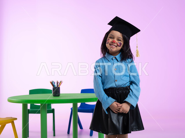 إيماءات وجه تدل على الحماس والسعادة بالنجاح في الروضة، الرسم بالألوان على وجوه الأطفال، رسومات وجه للبنات تدل على الأنوثة واللطافة، بورتريه لطفلة عربية خليجية سعودية ترتدي الزي الكاجوال وقبعة التخرج، مفهوم اللعب والتسلية، خلفية وردية