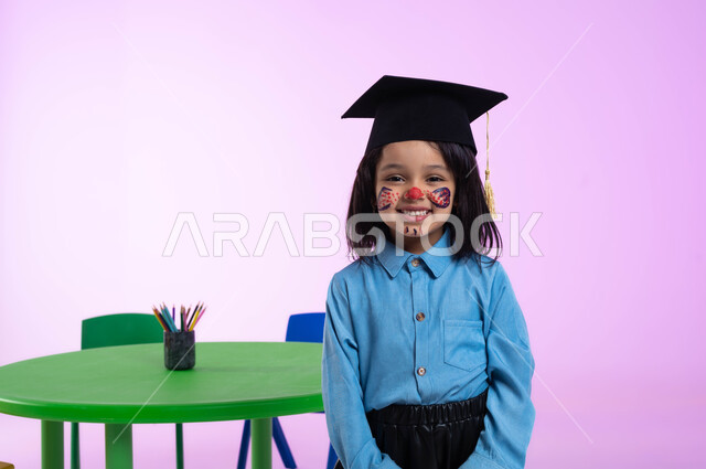 الرسم بالألوان على وجوه الأطفال، رسومات وجه للبنات تدل على الأنوثة واللطافة، بورتريه مقرب لطفلة عربية خليجية سعودية ترتدي الزي الكاجوال وقبعة التخرج، إيماءات وجه تدل على الحماس والسعادة بالنجاح في الروضة، مفهوم اللعب والتسلية، خلفية وردية