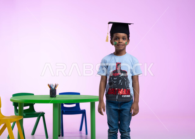 التلوين و الرسم بالوان صحية على وجوه الأطفال، إيماءات تدل على الثقة بالنفس، أشكال ورسومات وجه للأولاد تدل على اللطافة، بورتريه لطفل عربي خليجي سعودي يرتدي الزي الكاجوال وقبعة التخرج من الروضة، مفهوم اللعب و المرح، خلفية وردية