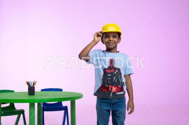 تعريف الأطفال على مفهوم الهندسة و المهن المستقبلية، دمج التعليم باللعب في الروضة، بورتريه  لفتى عربي خليجي سعودي مبتسم يرتدي الزي الكاجوال وخوذة الوقاية الخاصة بالعمل ينظر إلى الكاميرا بإيماءات الثقة، مهنة ووظيفة هندسية، خلفية زهرية