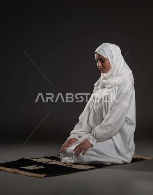 مفهوم أركان الاسلام والأيمان، الحفاظ على إقامة الصلوات الخمس بوقتها، التقرب الى الله تعالى بالإجتهاد في العبادة، بورتريه لإمرأة عربية سعودية خليجية ترتدي الحجاب والعباءة البيضاء تجلس على السجادة تقوم بأداء فريضة الصلاة، خلفية سوداء