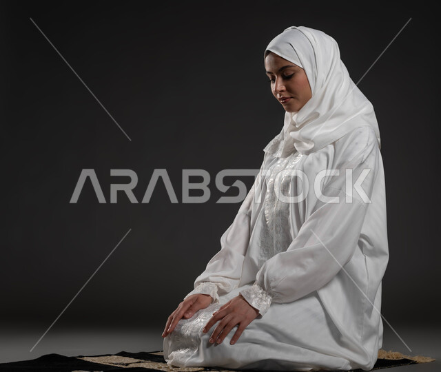 الحفاظ على إقامة الصلوات الخمس بوقتها، التقرب الى الله تعالى بالإجتهاد في العبادة، بورتريه لإمرأة عربية سعودية خليجية ترتدي الحجاب والعباءة البيضاء تجلس على السجادة تقوم بأداء فريضة الصلاة، مفهوم أركان الاسلام والأيمان، خلفية سوداء