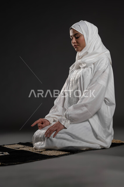 التقرب الى الله بالإجتهاد في العبادة والحفاظ على الصلوات، بورتريه لإمرأة عربية سعودية خليجية ترتدي الحجاب والعباءة البيضاء تجلس على سجادة الصلاة تقوم بالتشهد، رفع اصبع السبابة للأعلى، مفهوم أركان الاسلام والأيمان، الالتزام بأداء الفروض على وقتها, خلفية سوداء