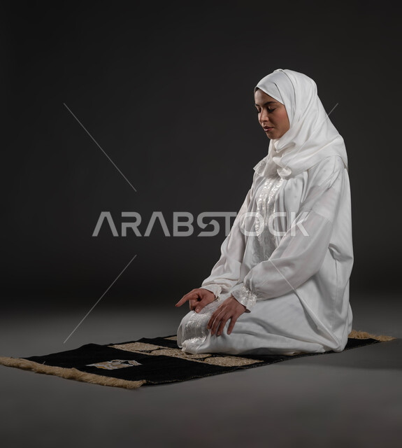 مفهوم أركان الاسلام والأيمان، الالتزام بأداء الفروض على وقتها, التقرب الى الله بالإجتهاد في العبادة والحفاظ على الصلوات، بورتريه لإمرأة عربية سعودية خليجية ترتدي الحجاب والعباءة البيضاء تجلس على سجادة الصلاة تقوم بالتشهد، رفع اصبع السبابة للأعلى، خلفية سوداء