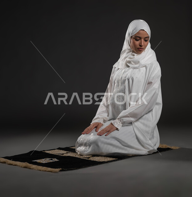 التقرب الى الله بالإجتهاد في العبادة والحفاظ على الصلوات، بورتريه لإمرأة عربية سعودية خليجية ترتدي الحجاب والعباءة البيضاء تجلس على سجادة الصلاة تقوم بالتسليم، الالتزام بأداء الفروض على وقتها, خلفية سوداء