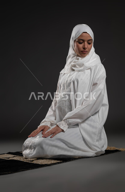 Commitment to performing the obligations on time, getting closer to God through diligence in worship and observing prayers, a portrait of a Saudi Gulf Arab woman wearing the hijab and white abaya sitting on the prayer rug making the salutations, black background.