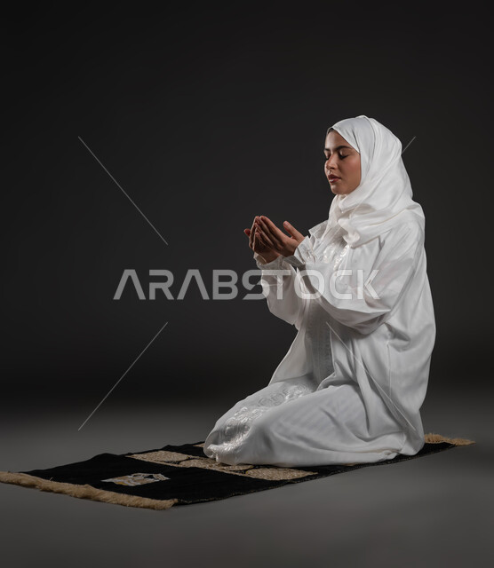 تعابير تدل على التوسل والتضرع، أداء الفروض على وقتها, التقرب الى الله من خلال الإجتهاد في العبادة والحفاظ على الصلوات، بورتريه لإمرأة عربية سعودية خليجية ترتدي الحجاب والعباءة البيضاء تجلس على سجادة الصلاة ترفع يديها وتدعو الله، خلفية سوداء