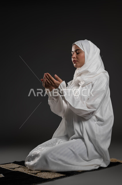 الطلب والتمني من الله، ايماءات تدل على الخشوع والتضرع، الإجتهاد في العبادة والحفاظ على الصلوات، بورتريه لإمرأة عربية سعودية خليجية ترتدي الحجاب والعباءة البيضاء تجلس على سجادة الصلاة ترفع يديها وتقوم بالدعاء، خلفية سوداء