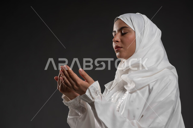 التمني والطلب من الله، ايماءات تدل على الخشوع والتضرع، الاجتهاد في الطاعات والأدعية والأذكار، العبادة والتقرب الى الله، بورتريه مقرب لإمرأة عربية خليجية سعودية ترتدي الحجاب والعباءة البيضاء ترفع يديها وتقوم بالدعاء، خلفية سوداء