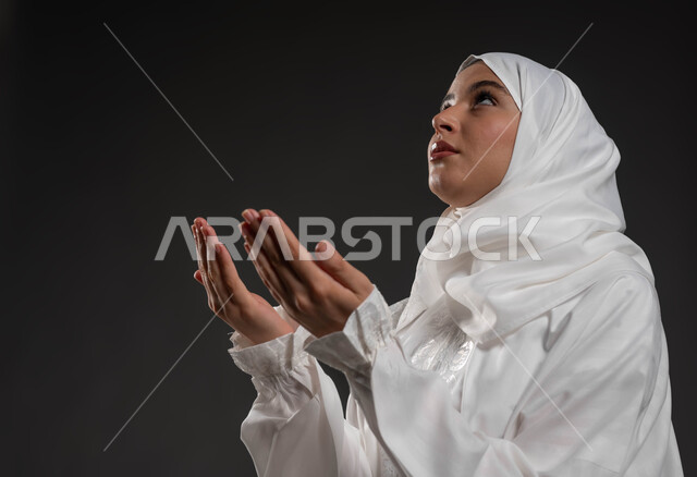 الطلب والتمني من الله، ايماءات تدل على الخشوع والتضرع، الاجتهاد في الطاعات والأدعية والأذكار، العبادة والتقرب الى الله، بورتريه مقرب لإمرأة عربية خليجية سعودية ترتدي الحجاب والعباءة البيضاء ترفع يديها وتقوم بالدعاء، خلفية سوداء