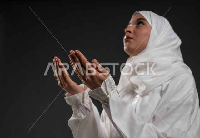 Diligence in obedience, supplications and remembrances, requesting and wishing from God, gestures indicating humility and supplication, worship and closeness to God, close-up portrait of a Saudi Gulf Arab woman wearing the hijab and white abaya, raising her hands and praying, black background.