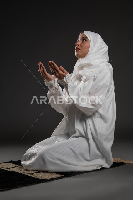 الدعاء والطلب والتمني من الله، ايماءات تدل على التوسل والتضرع، الإجتهاد في العبادة والحفاظ على الصلوات، بورتريه لإمرأة عربية سعودية خليجية ترتدي الحجاب والعباءة البيضاء تجلس على سجادة الصلاة ترفع يديها وتدعو الله، خلفية سوداء