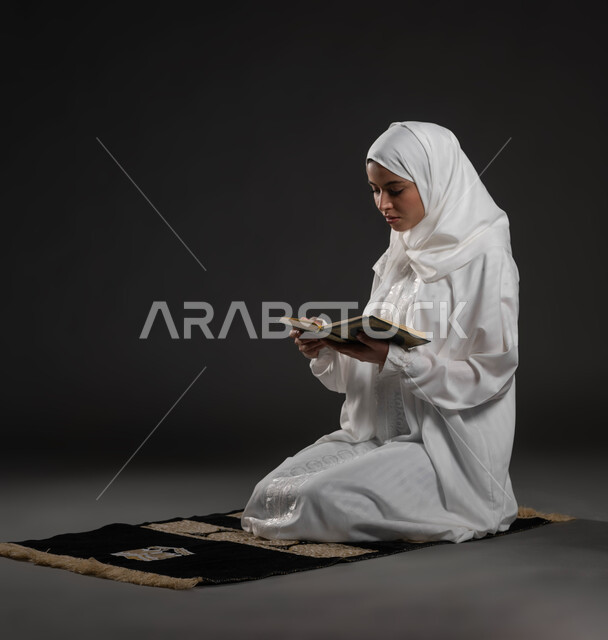 Getting closer to the Lord of the Worlds in the blessed month of Ramadan, recitation and reading of the Book of God Almighty, diligence and reverence in worship, portrait of a Saudi Arabian Gulf woman wearing the hijab and white abaya, holding the Holy Qur’an in her hand, sitting on the prayer rug, gray background